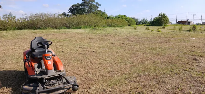 Trator rider laranja realizando a roçagem de uma grande área aberta, com árvores e construções ao fundo.