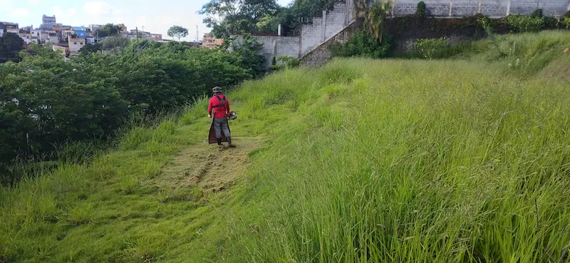Profissional com uniforme e roçadeira costal limpando um terreno inclinado, com casas ao fundo.